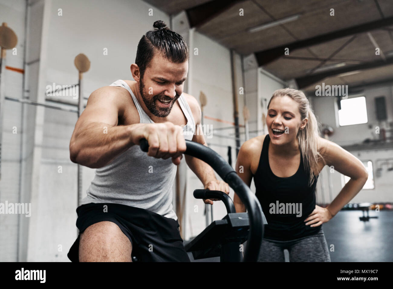 Smiling young woman cheering on her male friend riding a stationary bike during a workout session together at the gym Stock Photo