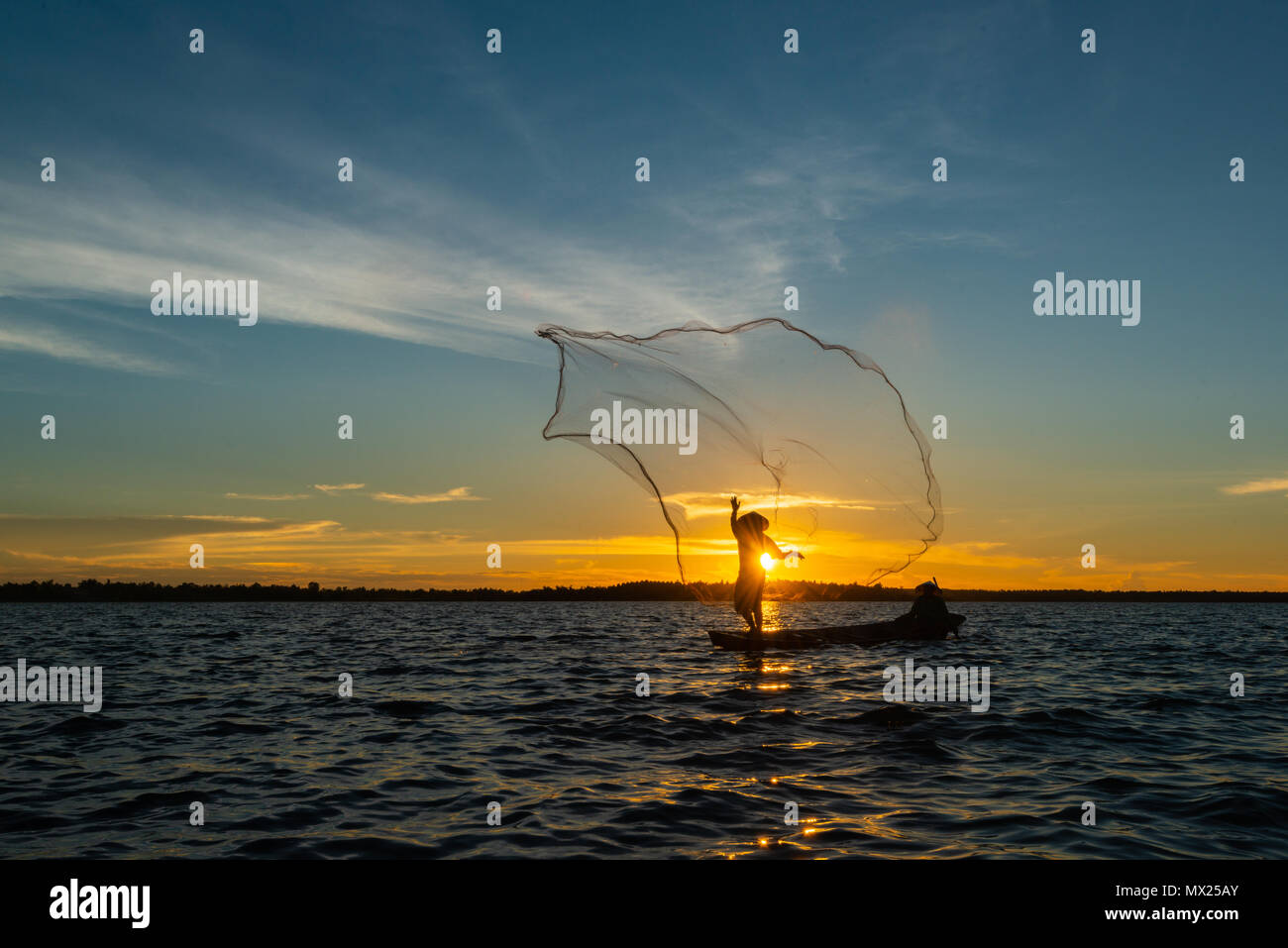 Un-identified silhouette fisher man on boat fishing by throwing fishing net to river during sunset in rural of Thailand Stock Photo