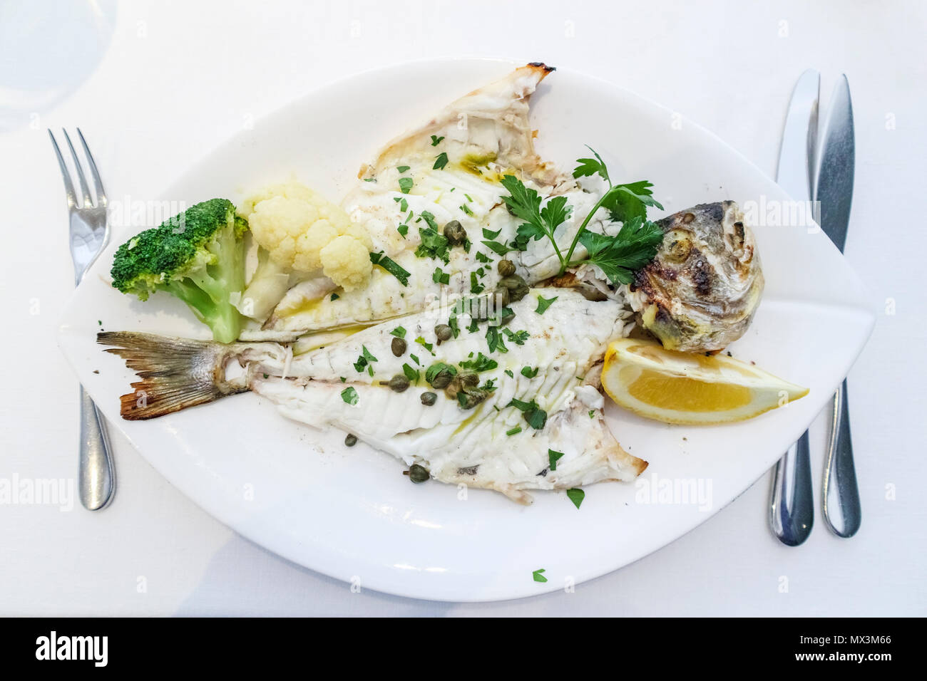 Fine dining: a main course of grilled sea bream (dorade) served on a white plate with a garnish of capers, broccoli and cauliflower and slice of lemon Stock Photo