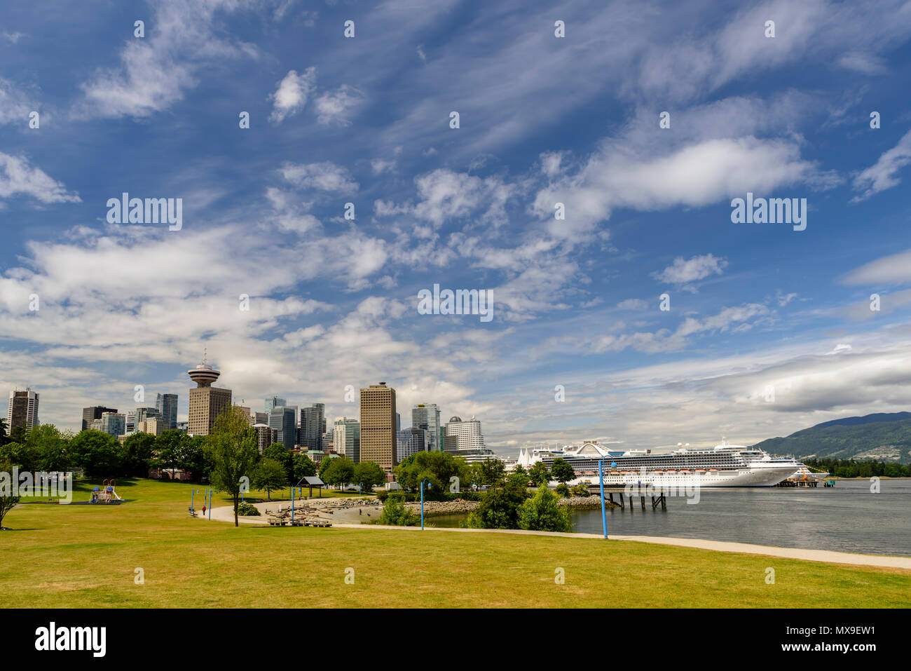 A summer day in a megalopolis city park with skyscrapers, a blue sky with clouds, a white ship, a blue ocean, mountains and green trees Stock Photo