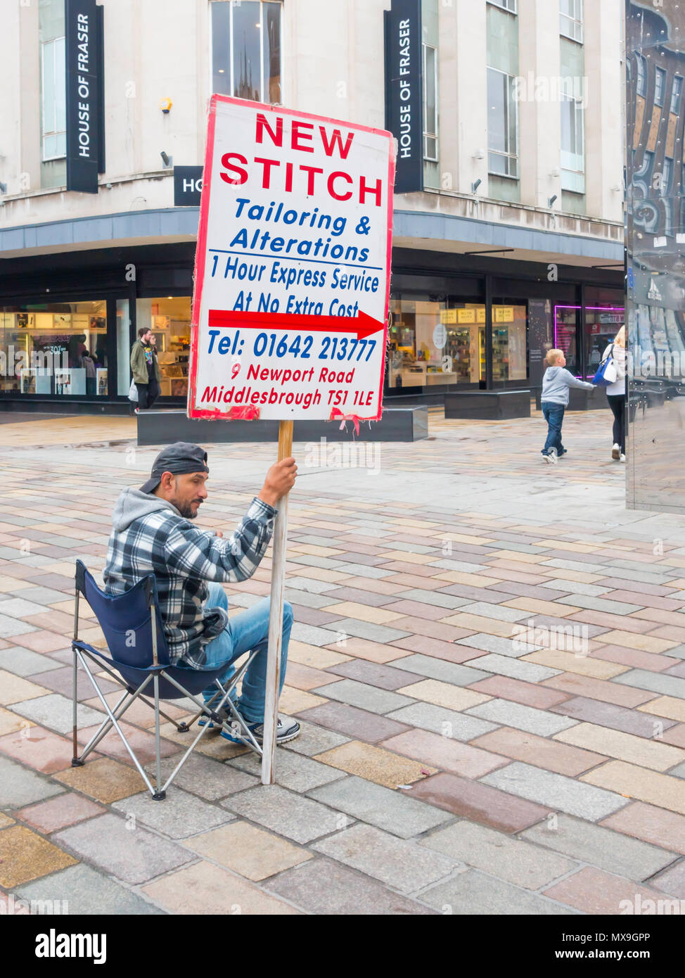 East Asian man seated at a pedestrianised junction in Middlesbrough Town Centre holding an advertising placard for New Stitch a Turkish Tailor shop Stock Photo