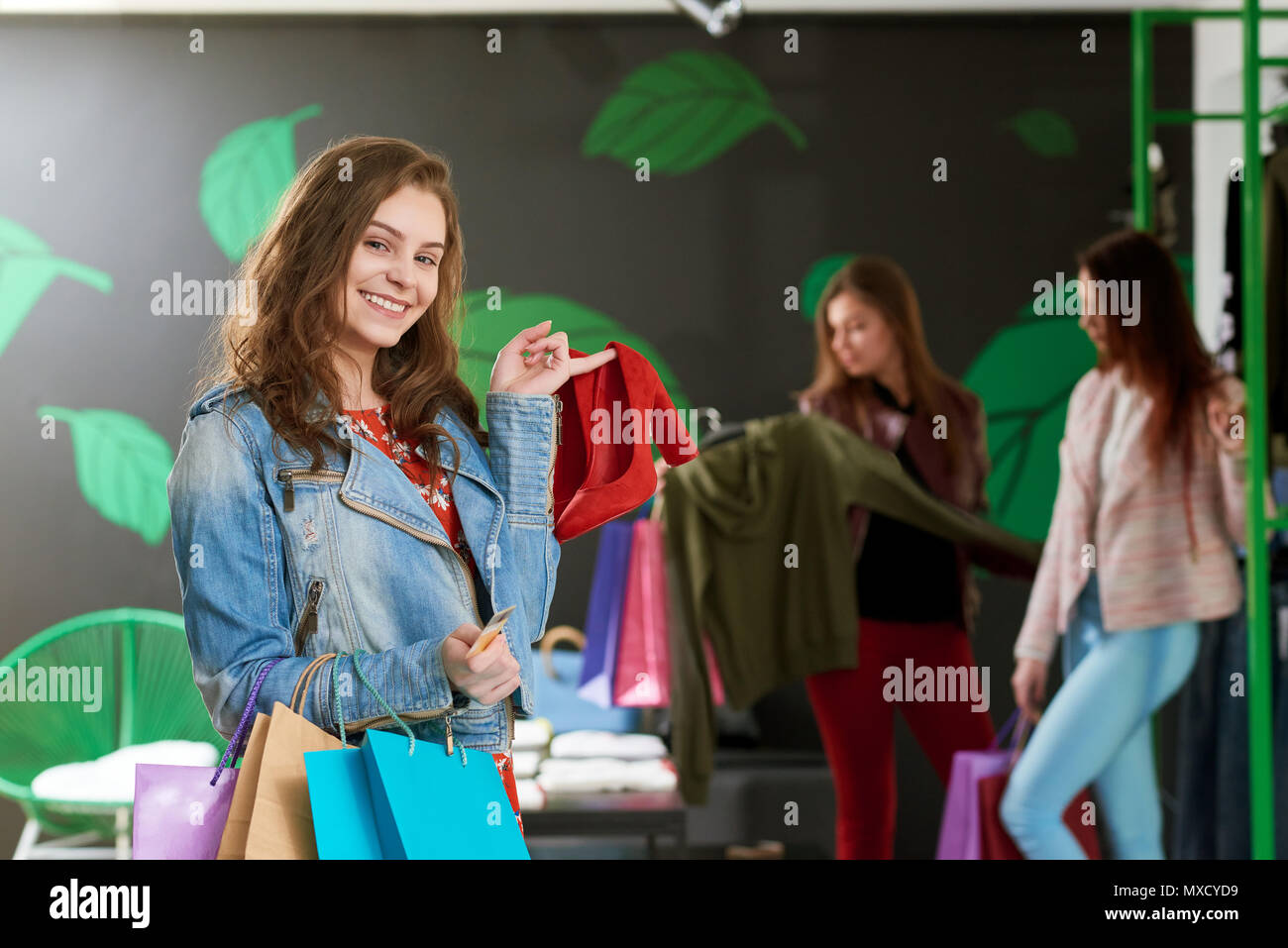 Cropped photo of nice girl holding pair of red high heel shoes in the shop. Keeping colorful packages and golden credit card in another hand. Two young friends on background choothing clothes. Stock Photo