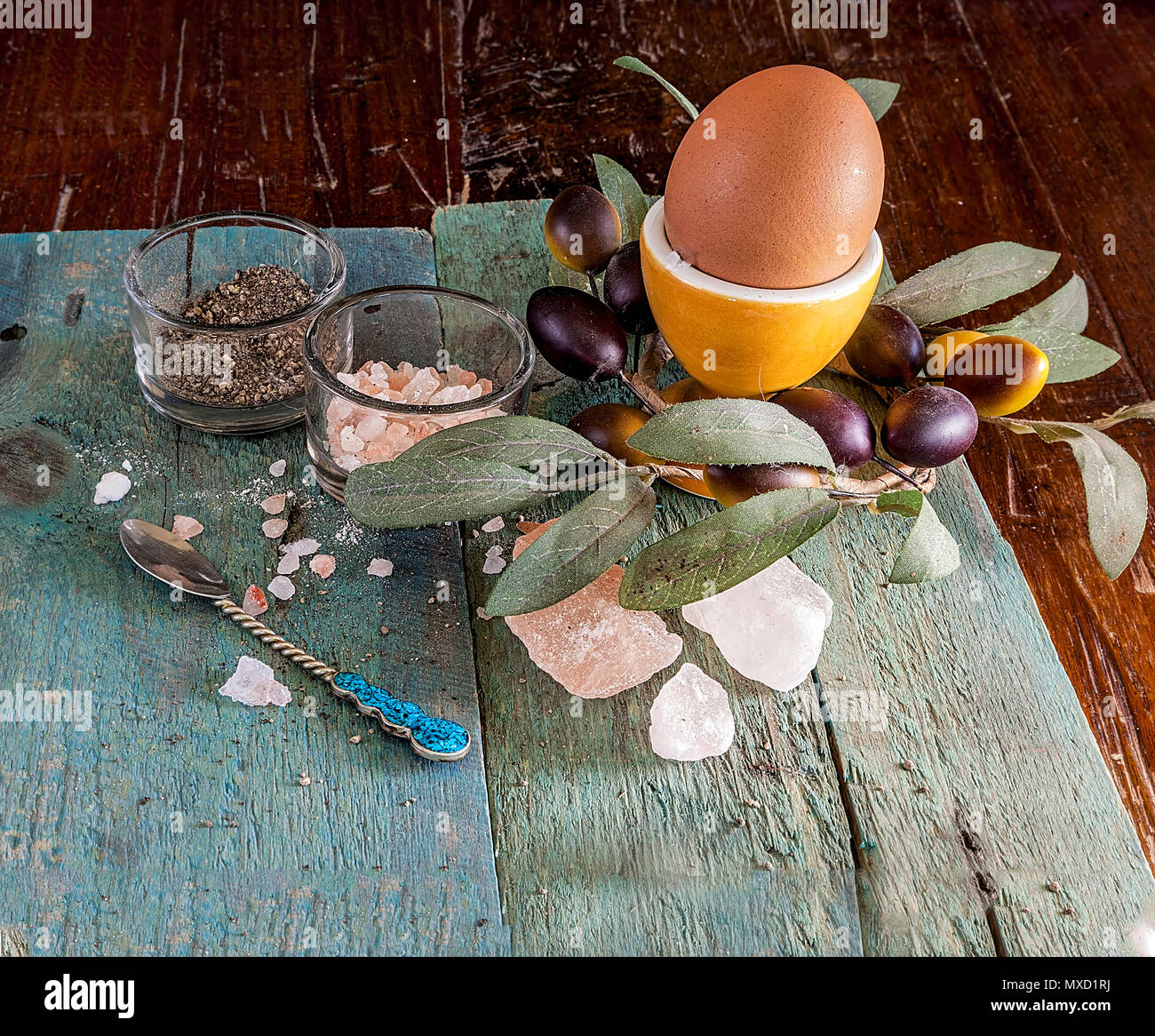 Single egg in a egg cup on a wooden surface with an assortment of crystal Hymalain pink salt and pepper. Olive decorative reef in the background. Stock Photo