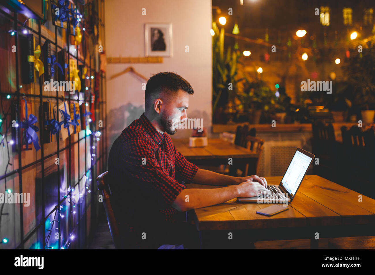 A young handsome Caucasian man with beard and toothy smile in a red checkered shirt is working behind a gray laptop sitting at a wooden table. Hands on the keyboard. In the evening at the coffee shop Stock Photo
