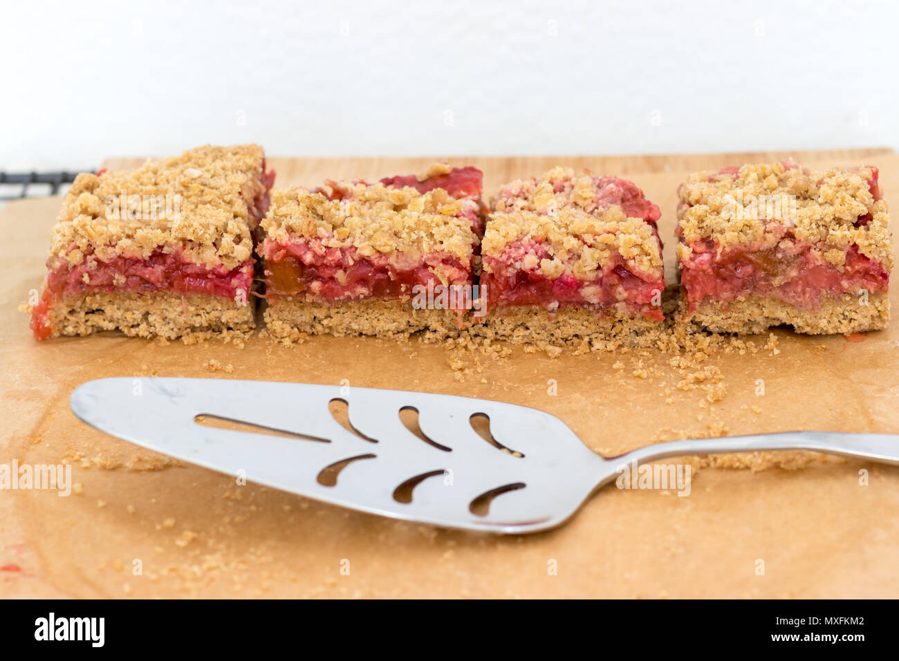 Slices of rhubarb & strawberry crumble bars, displayed on baking paper with a silver cake server. These fruity treats are a healthy snack or dessert. Stock Photo