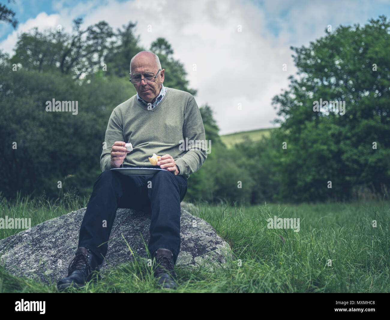 A senior man is sitting on a rock in nature and is having a picnic for ...