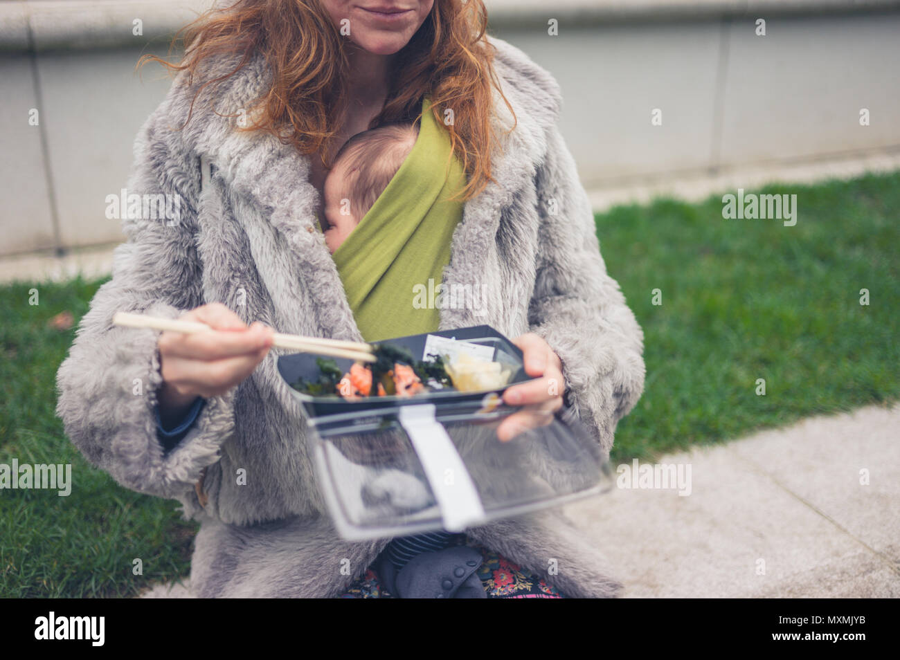 A young mother with a baby in a carrier sling is eating sushi outside Stock Photo