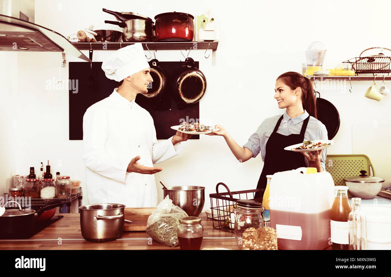 Portrait of diligent young smiling man cook giving to waitress ready to
