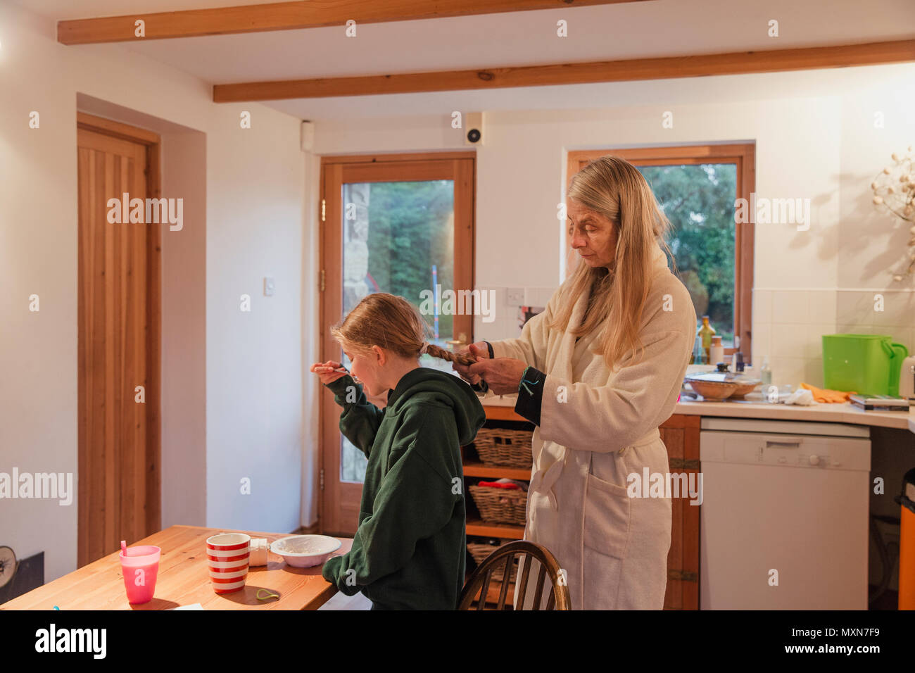Mother plaiting her daughters hair as she eats her breakfast in the kitchen. Stock Photo