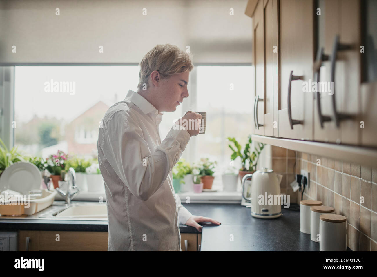 Side view of a young adult businessman standing at the kitchen counter blowing on a cup of tea to cool it down. Stock Photo