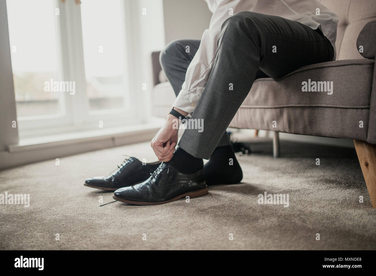 Unrecognisable person adult male sitting on a sofa in a domestic room tying his shoelaces. Stock Photo