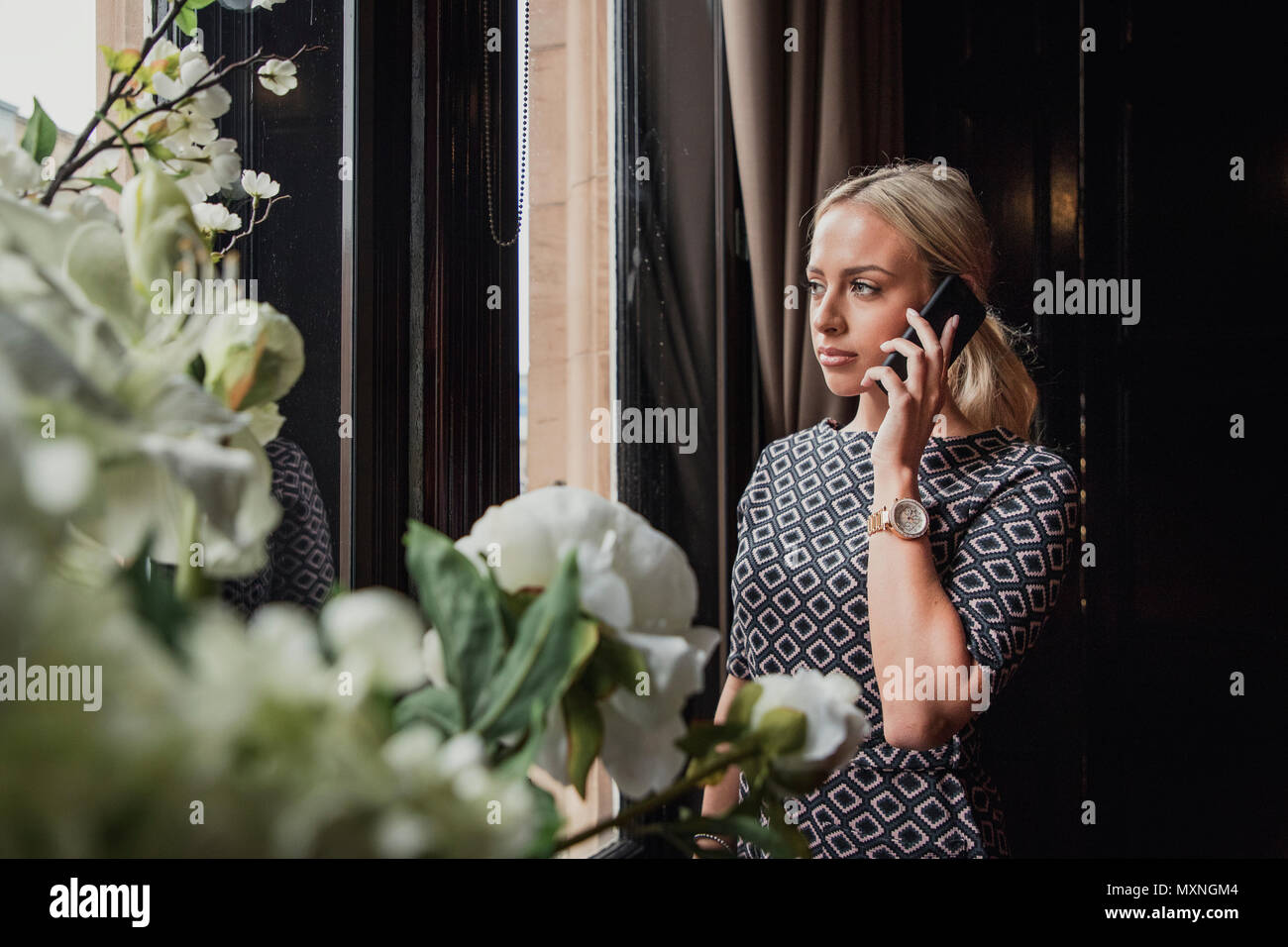 Young adult businesswoman standing looking out the window. She is making an important phone call at work and has a serious facial expression. Stock Photo