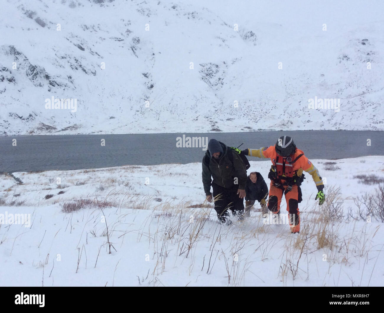 An air crew member of an MH-65 Dolphin detached to the Coast Guard Cutter Alex Haley assists two men who survived a helicopter crash near the Dutch Harbor Airport on Unalaska, Alaska, Dec. 2, 2016. Their helicopter reportedly encountered whiteout conditions and then crashed. U.S. Coast Guard photo by Lt. Craig Hermiller. Stock Photo