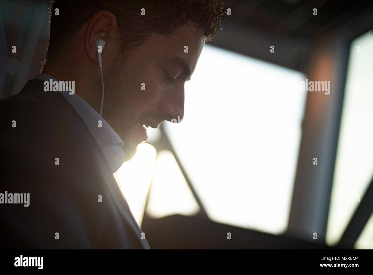 Side profile of an affluent well dressed young business man inside an office wearing ear buds in winter sunshine Stock Photo