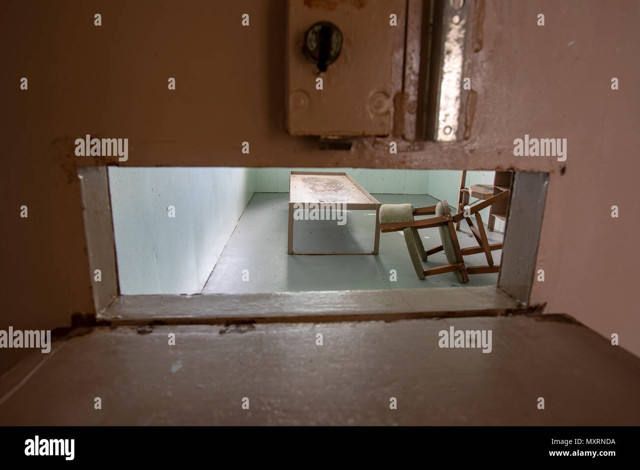 Metal bed frame with chair and desk inside solitary confinement cell through metal door slat in old prison. Stock Photo