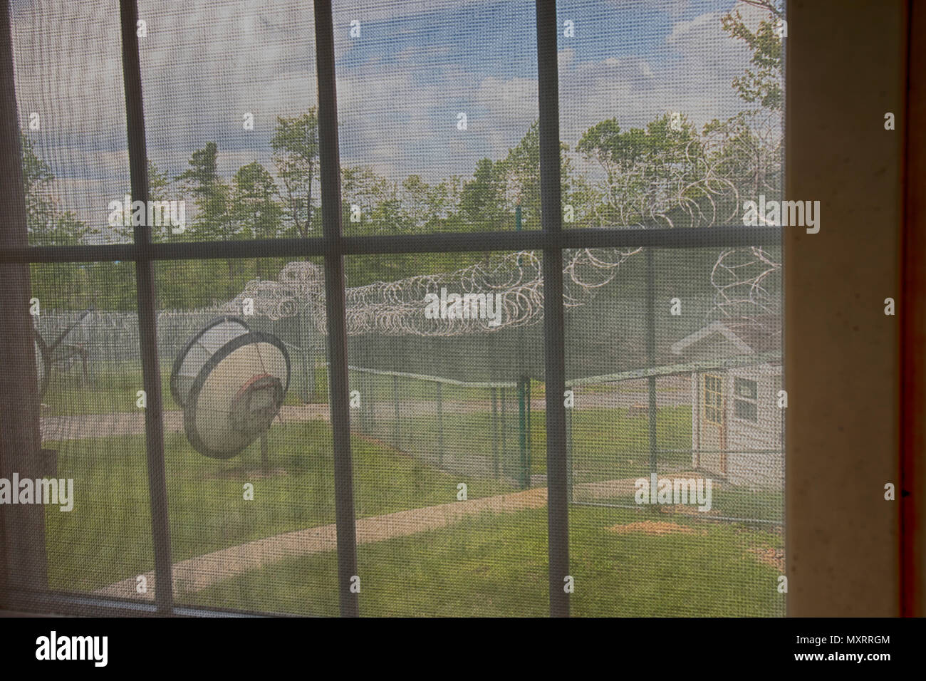 View through bars on window from inside abandoned prison thourgh mesh and bars looking at yard with guard house, metal fencing with razor wire and sat Stock Photo