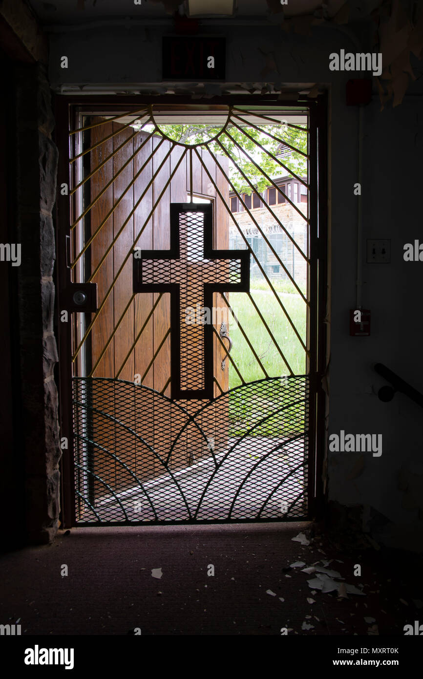 Light coming through metal gate with cross from inside church in prison yard with razor wire