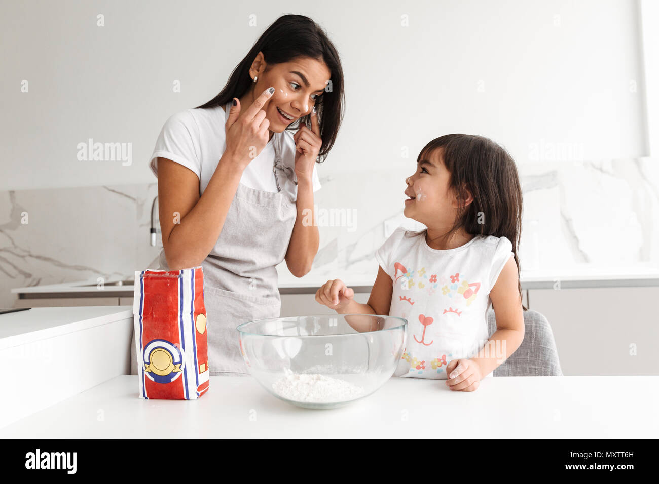 Happy brunette woman wearing apron cooking with her little daughter together in kitchen at home putting flour on face for fun Stock Photo