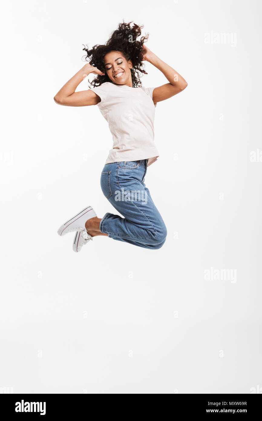 Full length portrait of adorable american woman with afro hairstyle wearing jeans and t-shirt jumping and rejoicing with perfect smile isolated over w Stock Photo