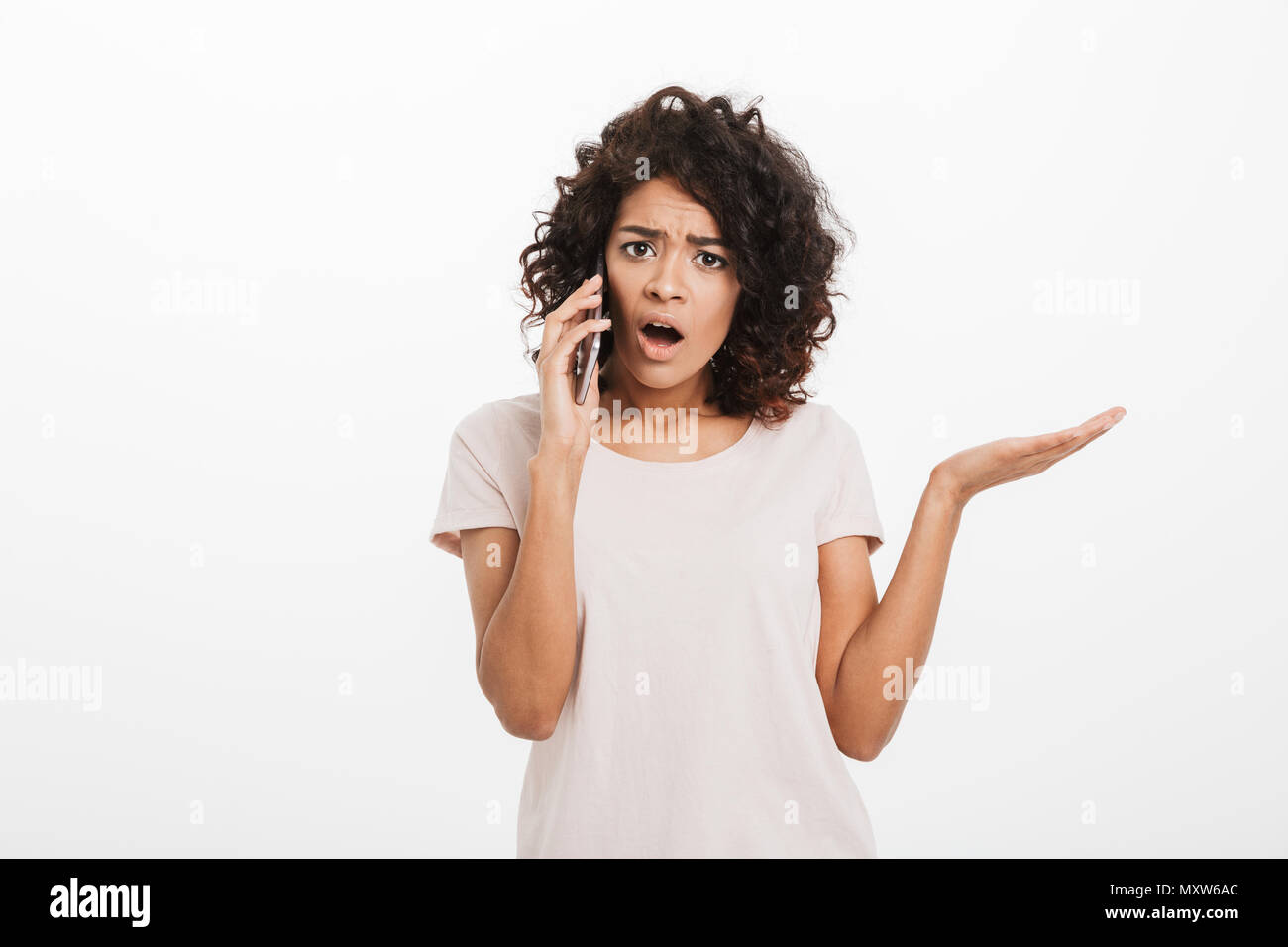 Portrait of serious puzzled woman with afro hairstyle wearing t-shirt having unpleasant conversation on cell phone isolated over white background Stock Photo