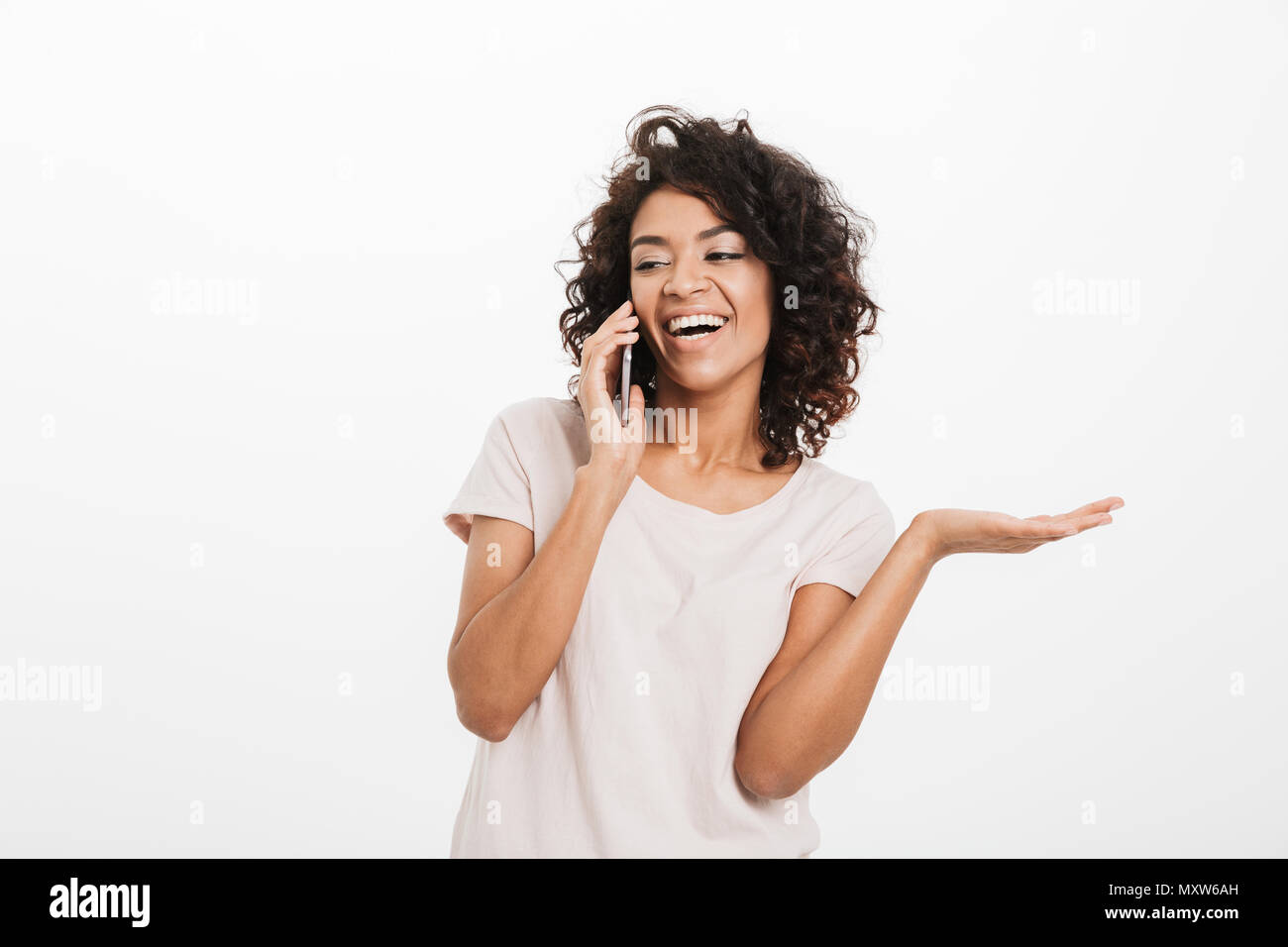 Happy american woman with afro hairstyle wearing t-shirt talking on smartphone and gesturing hand aside isolated over white background Stock Photo