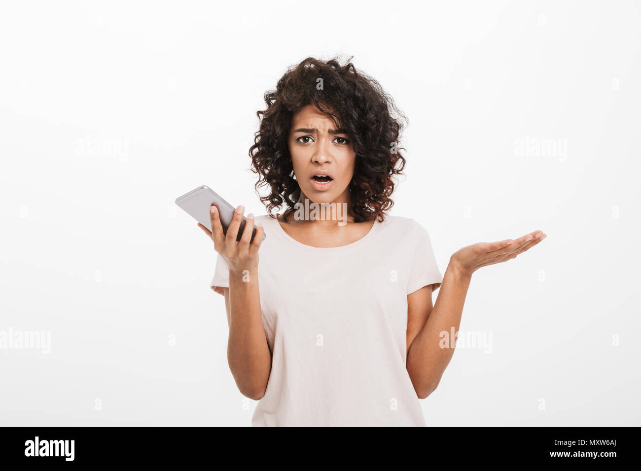 Portrait of confused puzzled woman with afro hairstyle wearing t-shirt holding cell phone and expressing misunderstanding isolated over white backgrou Stock Photo