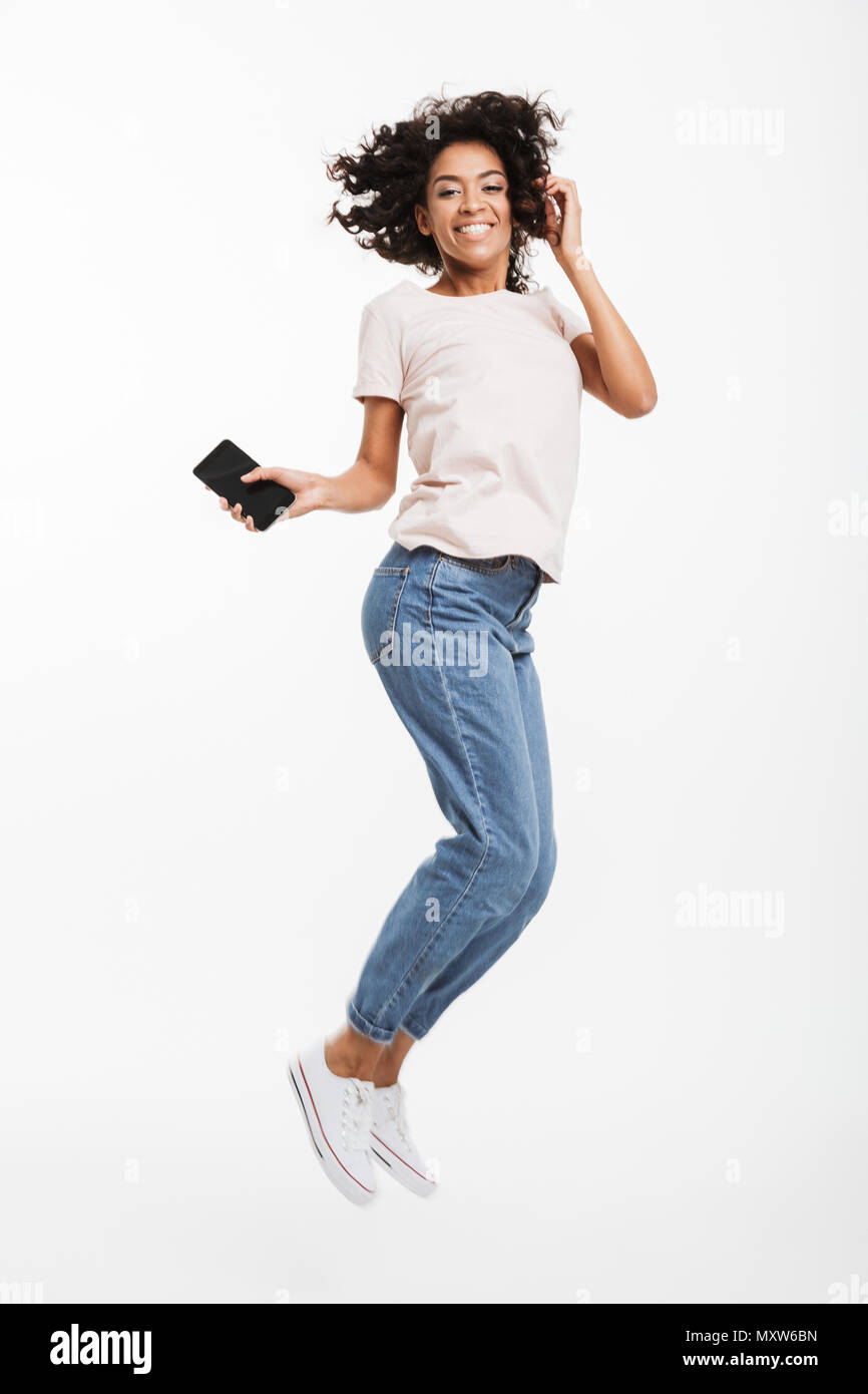 Full length image of happy african american woman with brown curly hair wearing jeans and t-shirt levitating with smartphone in hand isolated over whi Stock Photo