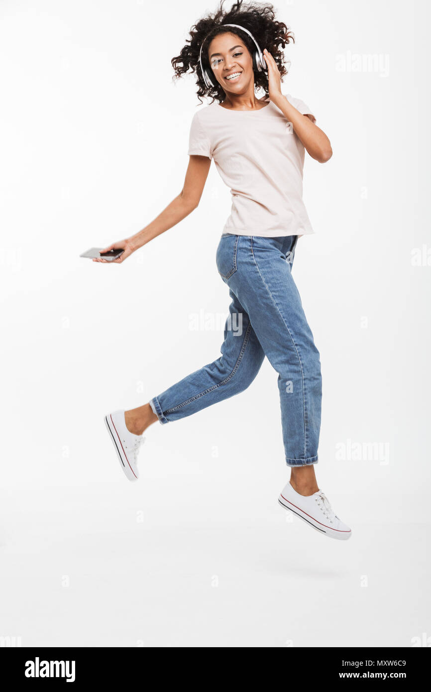 Full length photo of cheerful american woman with brown curly hair enjoying music listening via wireless headphones and mobile phone isolated over whi Stock Photo