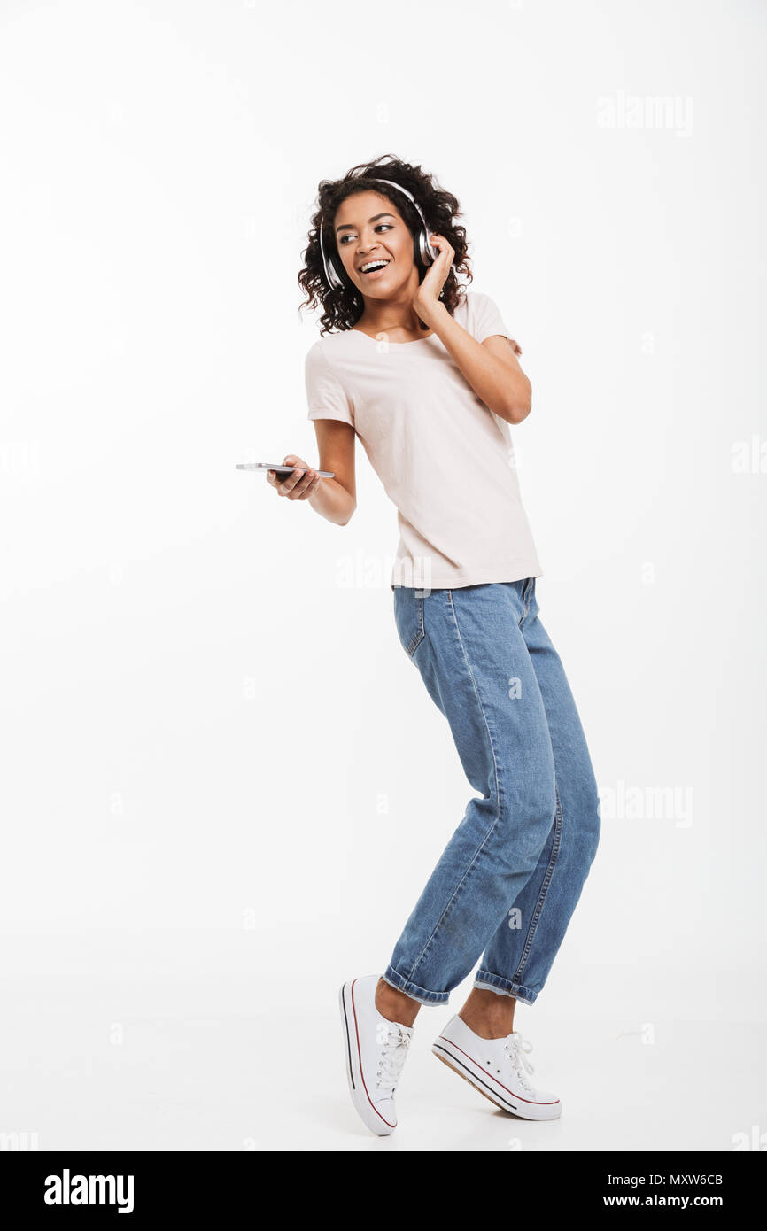 Full length image of cute african american woman with brown curly hair listening to music using wireless headphones and smartphone isolated over white Stock Photo