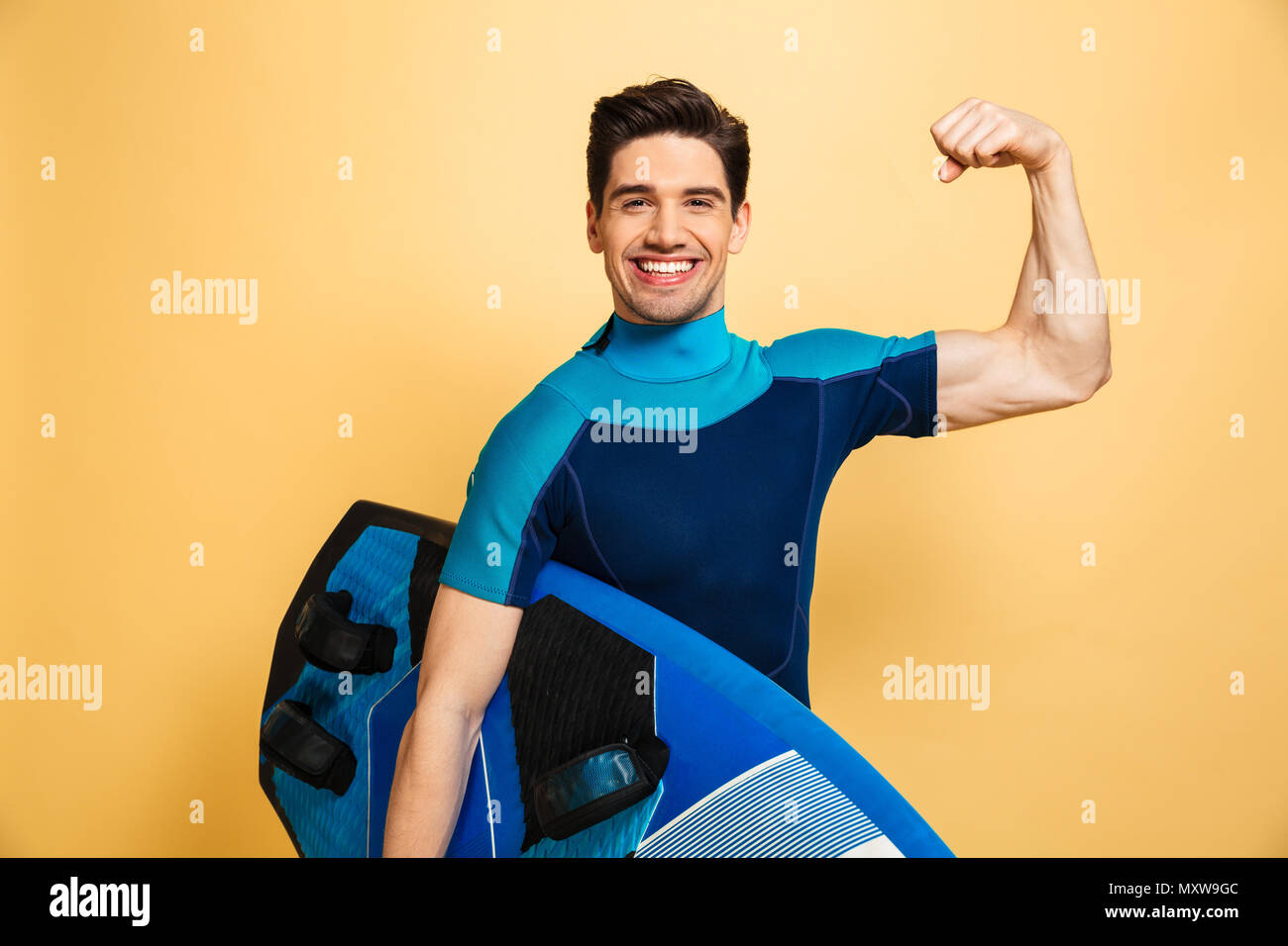 Portrait of a happy young man dressed in swimsuit flexing biceps while holding a surfboard isolated over yellow background Stock Photo