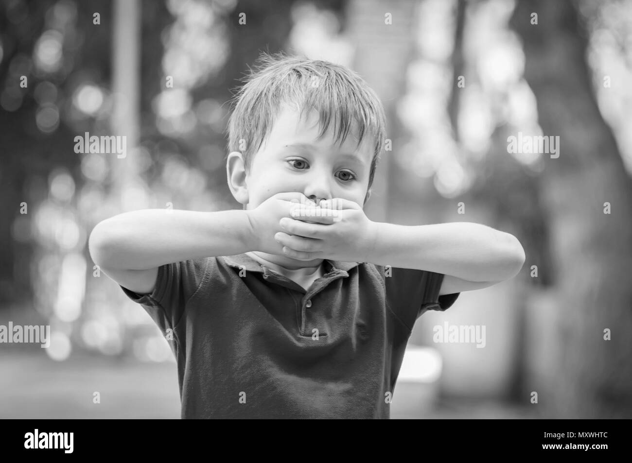 Serious little Caucasian boy closing his mouth with hands. Illustrative image for childhood trauma concept, child traumatic experience. Black white Stock Photo