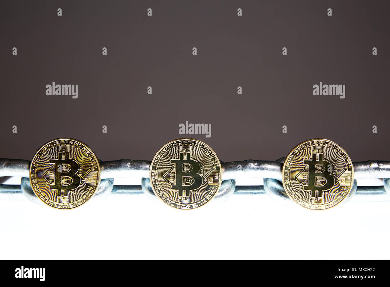 Three Bitcoins sits on a white glowing surface while a heavy steel metal chain sits behind.  Concepts of block chain technology and digital currency. Stock Photo