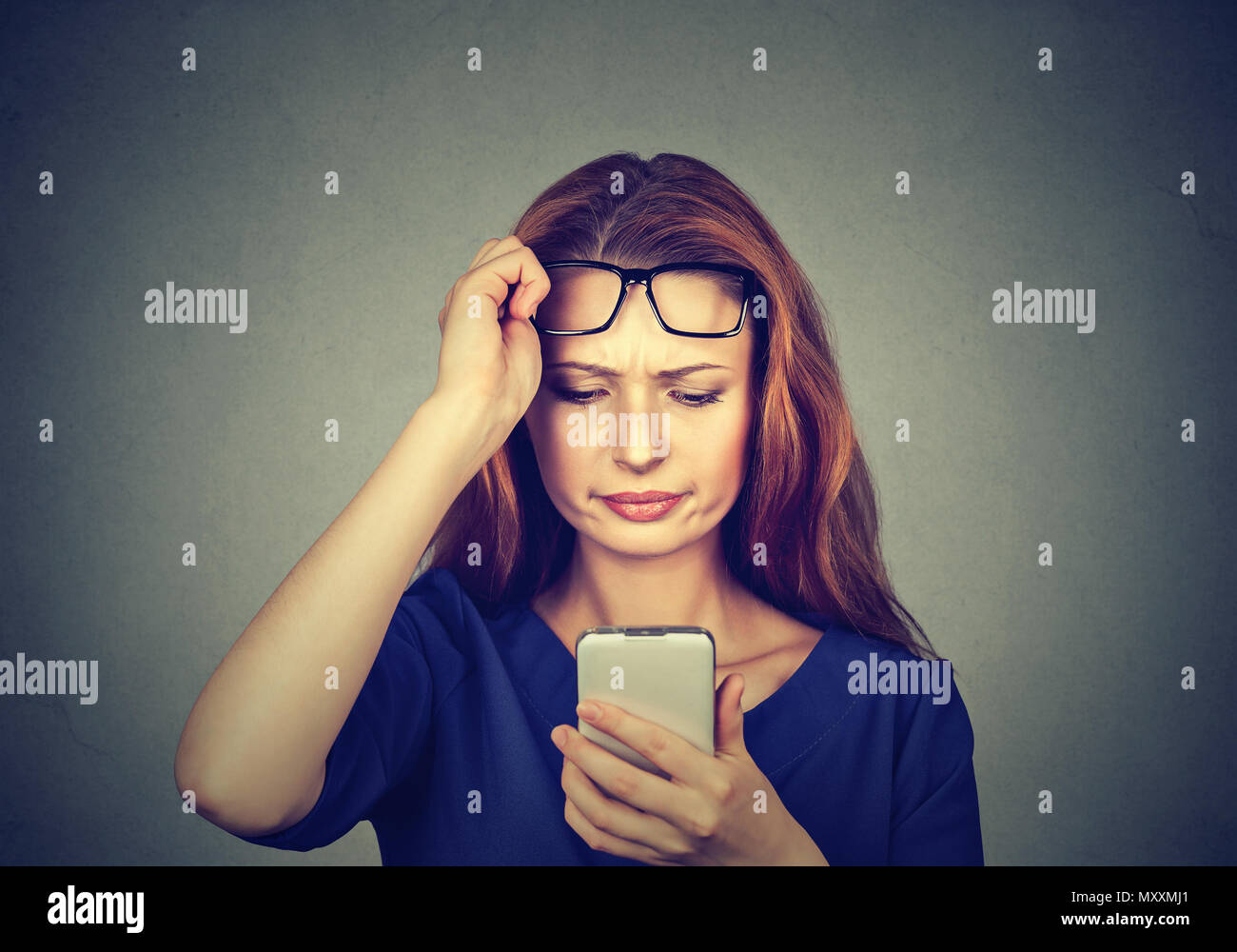 Young redhead girl holding eyeglasses up and watching smartphone having problems with eyes Stock Photo