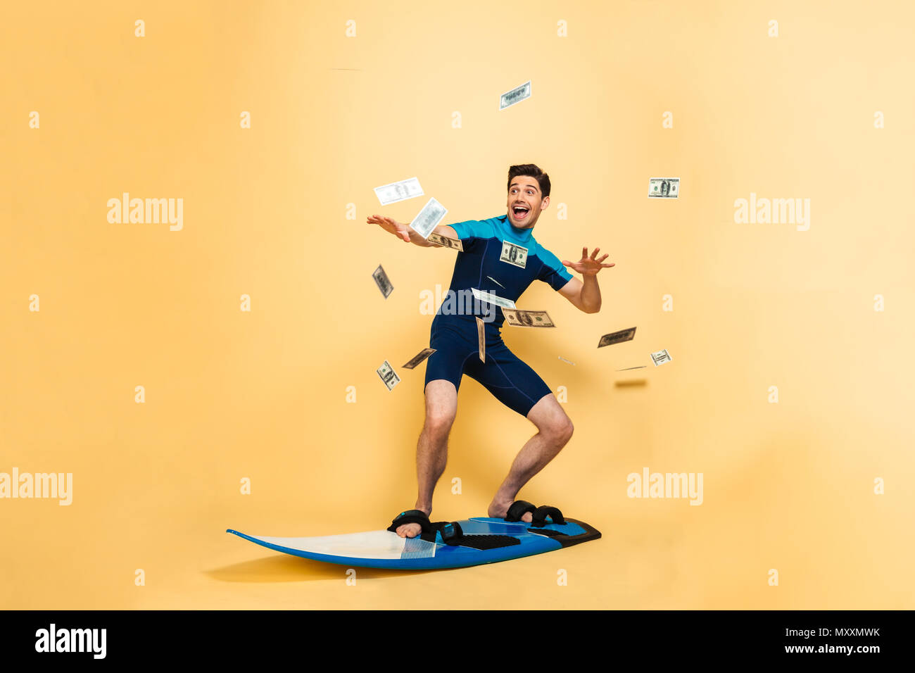 Full length portrait of a happy young man dressed in swimsuit surfing on a board under money shower isolated over yellow background Stock Photo