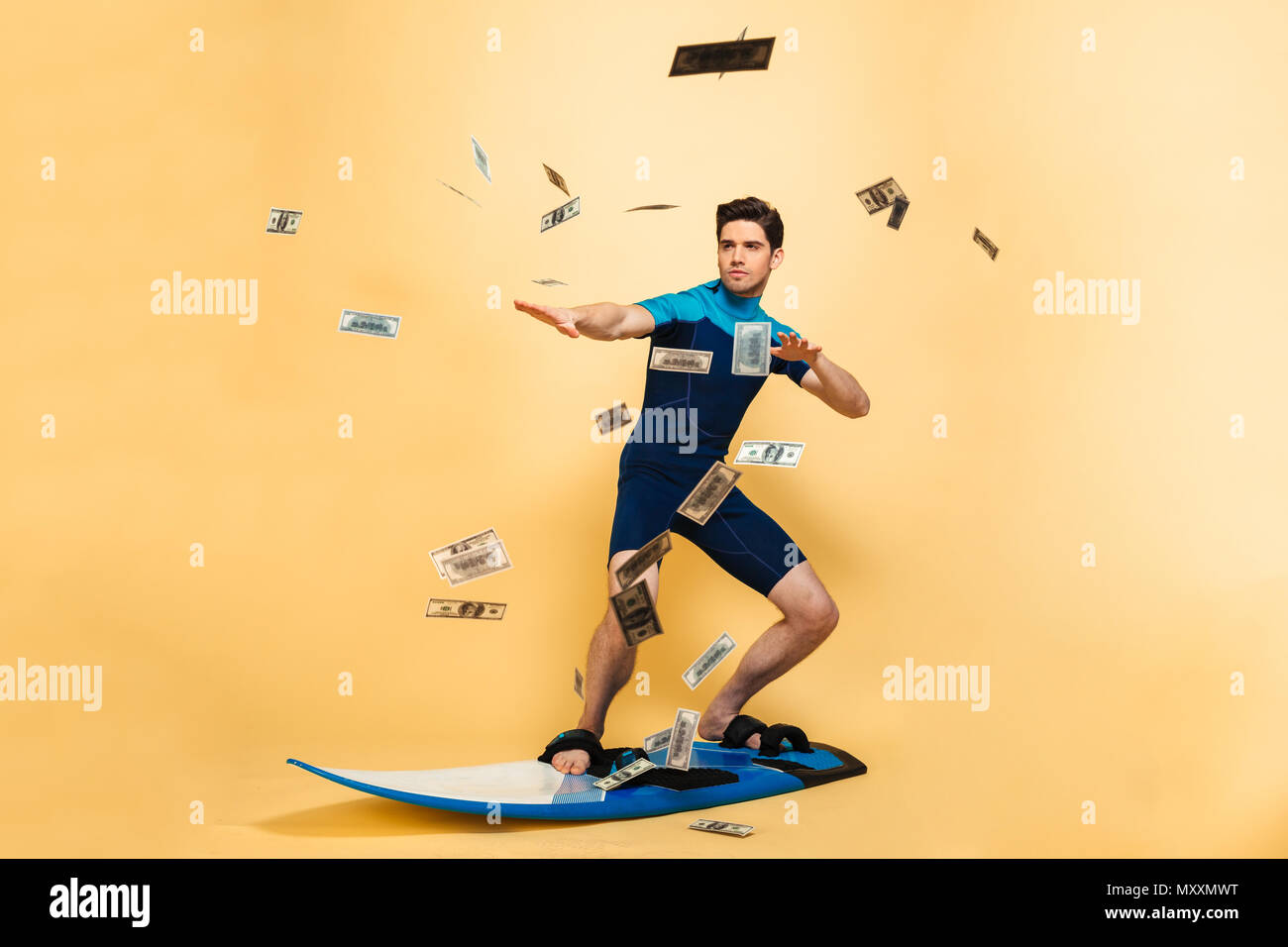 Full length portrait of a confident young man dressed in swimsuit surfing on a board under money shower isolated over yellow background Stock Photo