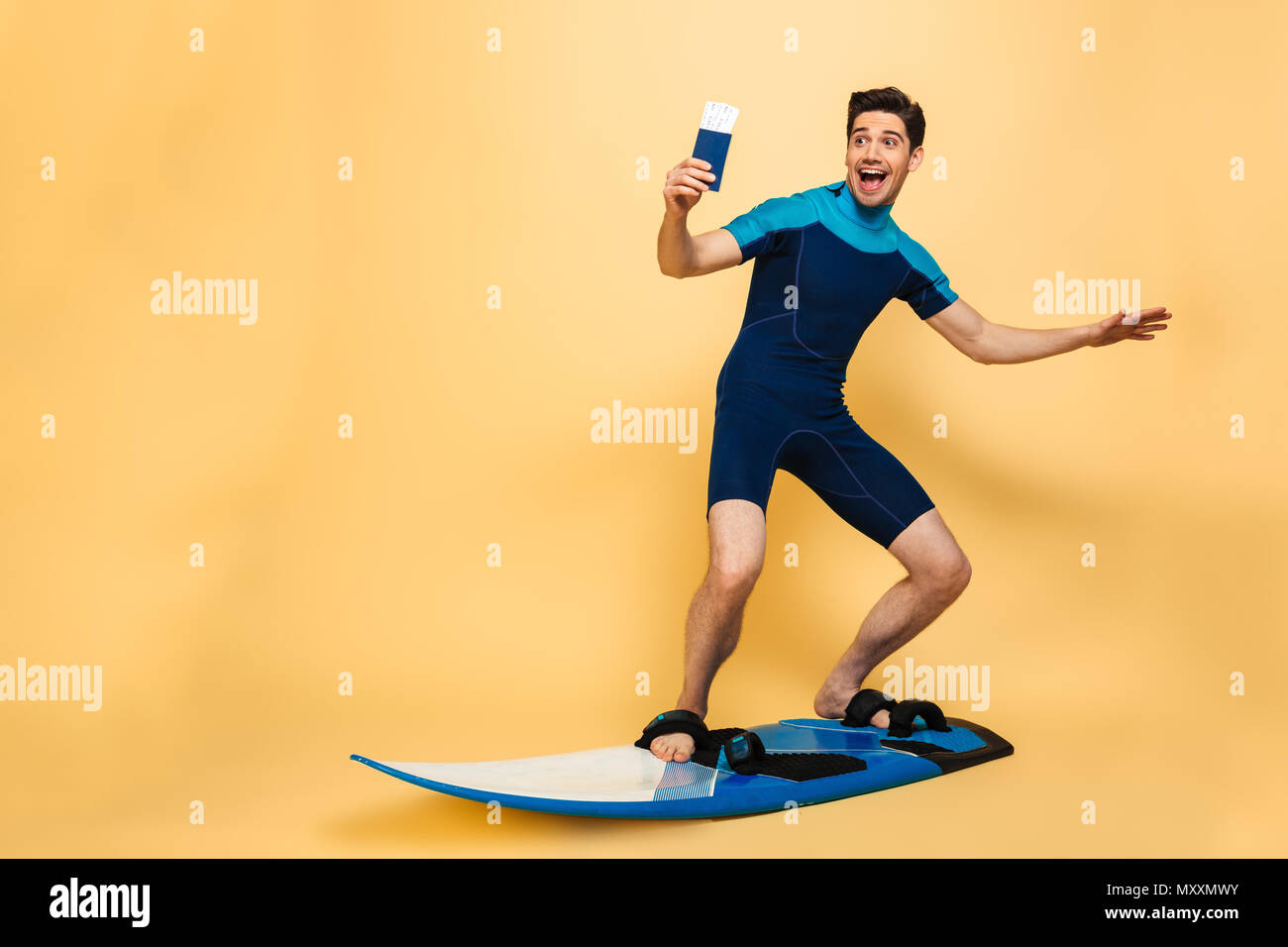 Full length portrait of a happy young man dressed in swimsuit holding passport while surfing on a board under money shower isolated over yellow backgr Stock Photo