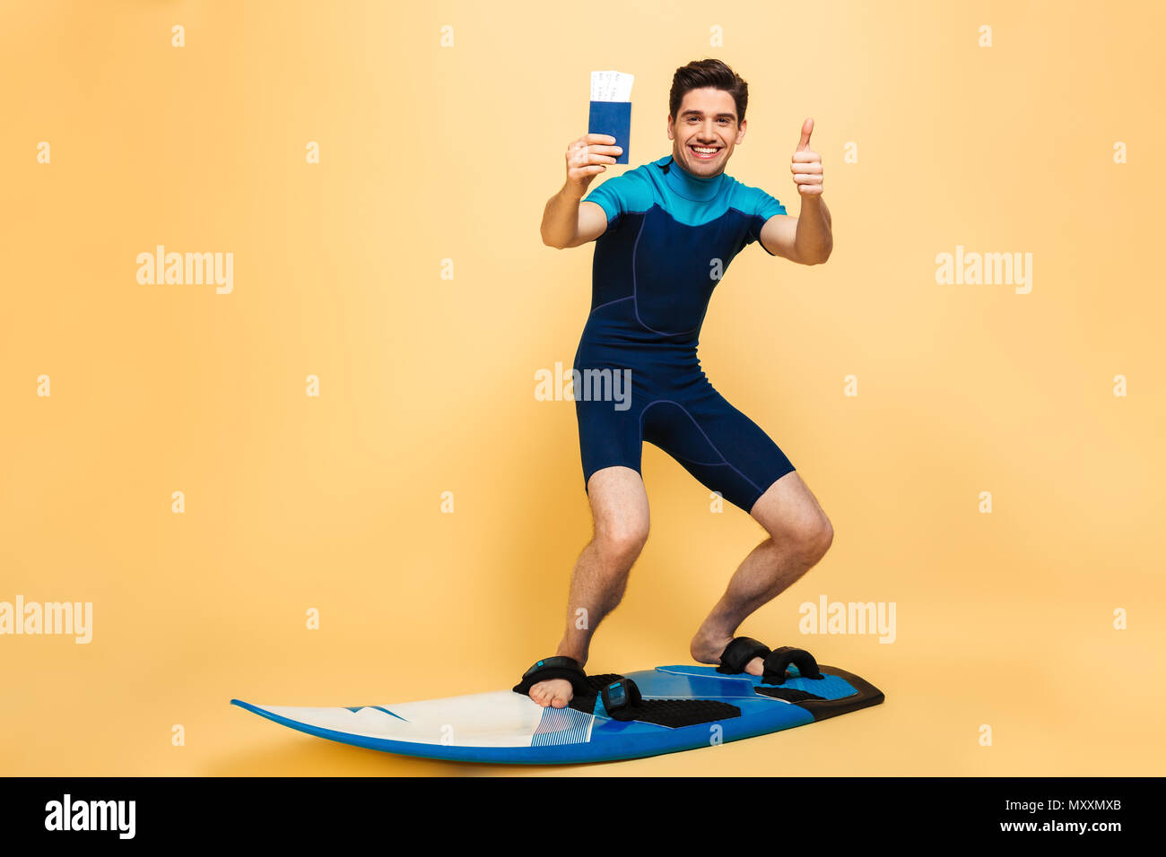 Full length portrait of a happy young man dressed in swimsuit showing passport while surfing on a board under money shower isolated over yellow backgr Stock Photo