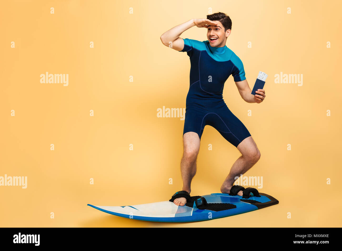 Full length portrait of an excited young man dressed in swimsuit showing passport while surfing on a board under money shower isolated over yellow bac Stock Photo