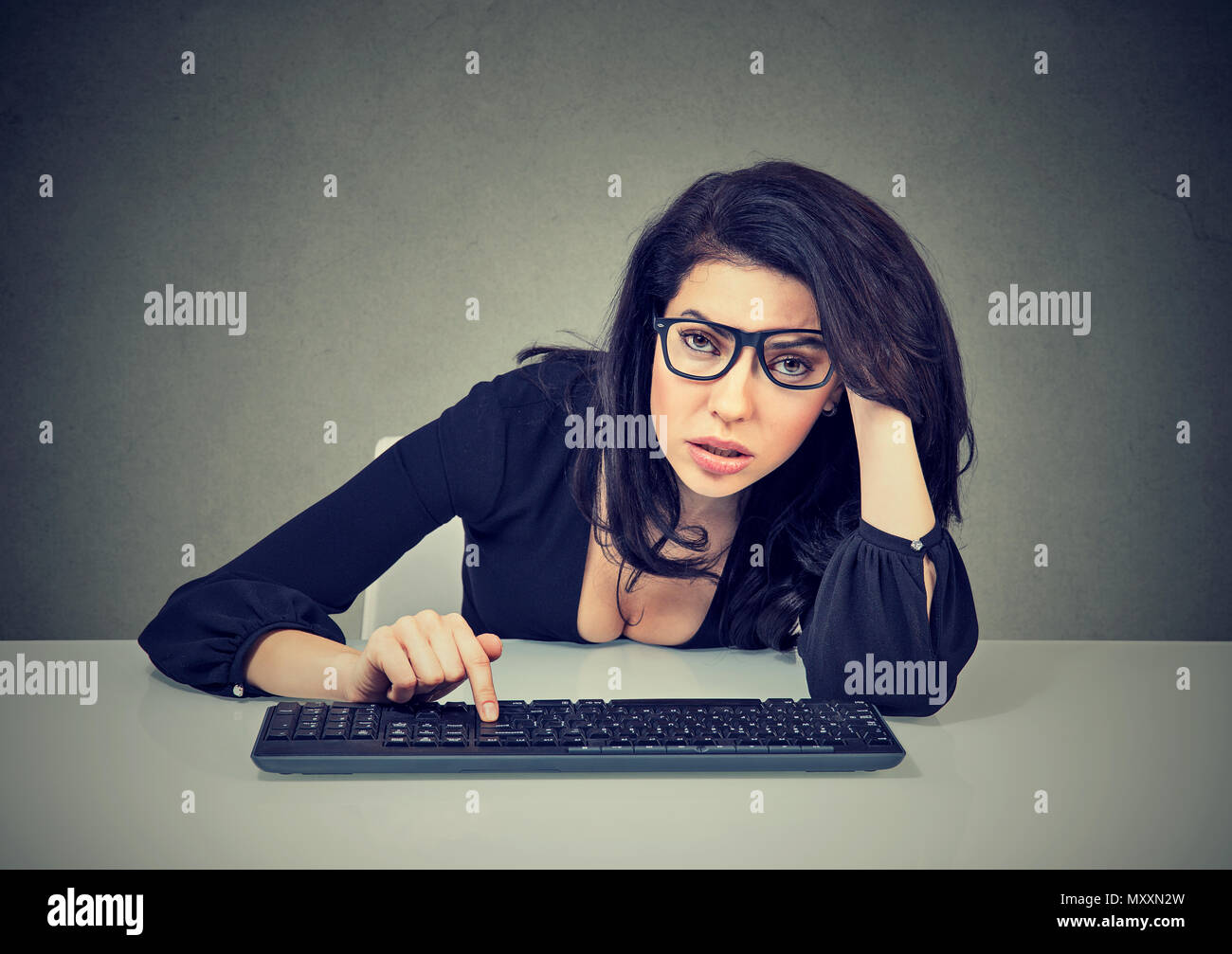 Tired and unmotivated young woman in glasses sitting lazily at table and typing on keyboard looking at camera Stock Photo