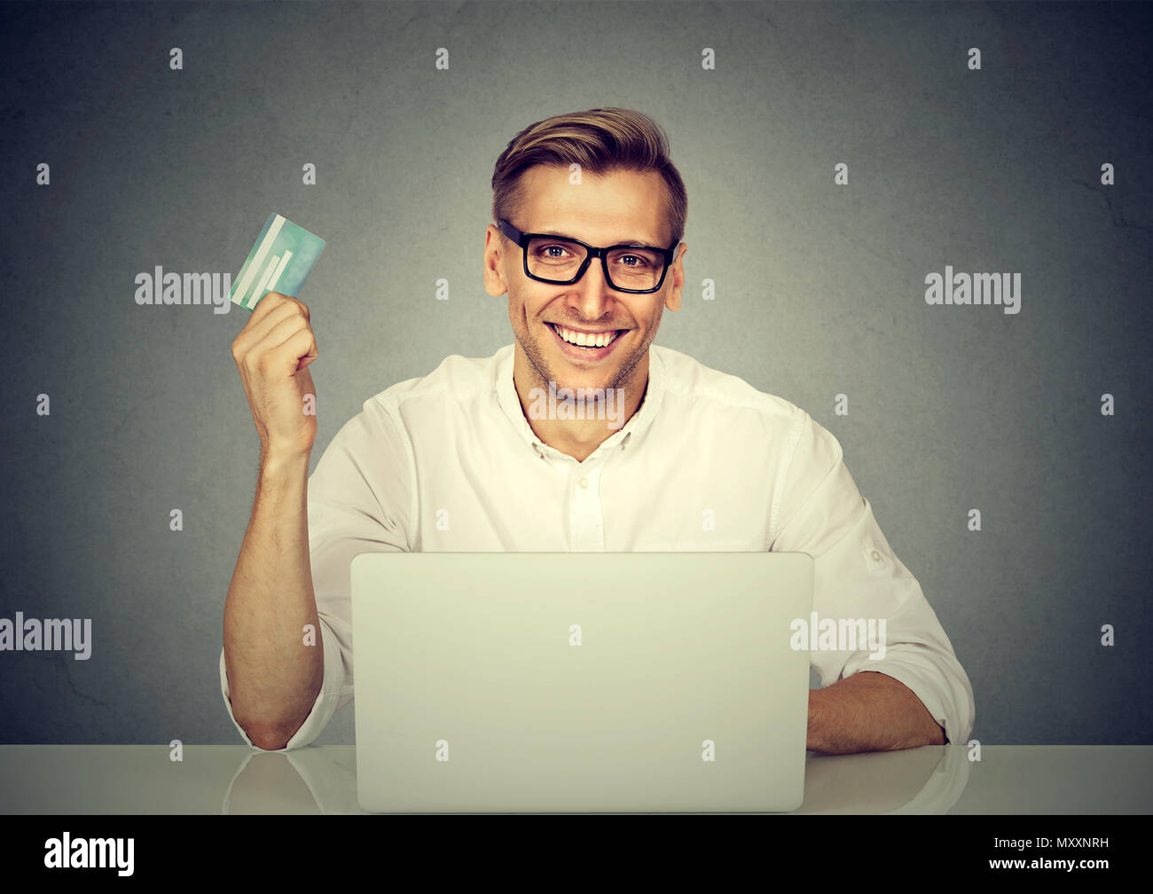 Stylish happy man in glasses sitting at table with laptop and doing purchases in Internet with credit card Stock Photo