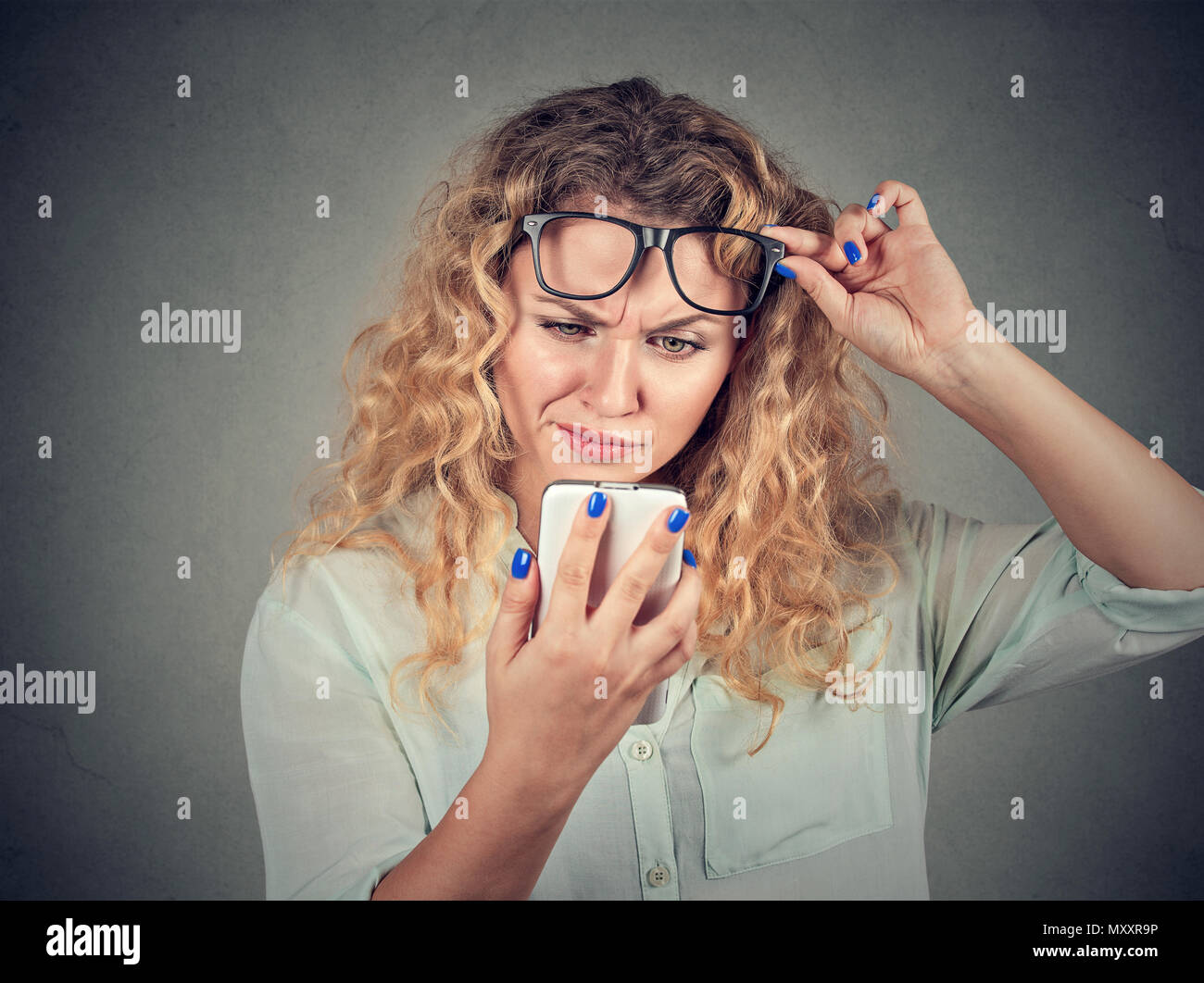 Pretty blond woman taking glasses up and exploring smartphone with doubt and confusion Stock Photo
