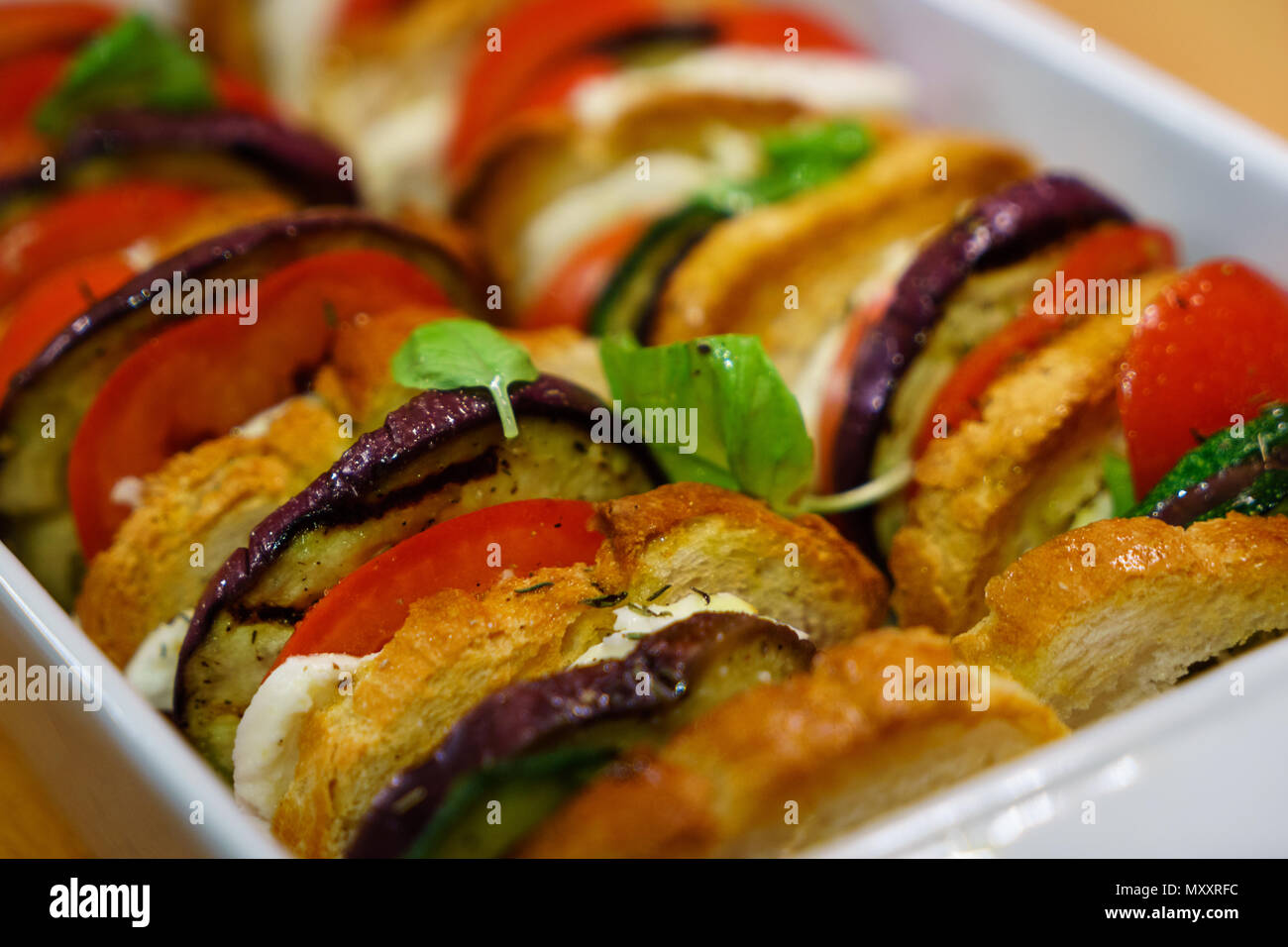 Flat lay top view of oven baked sliced vegetables (tomatoes, aubergines and courgettes) with mozzarella cheese and baguette slices dressed with basil. Stock Photo