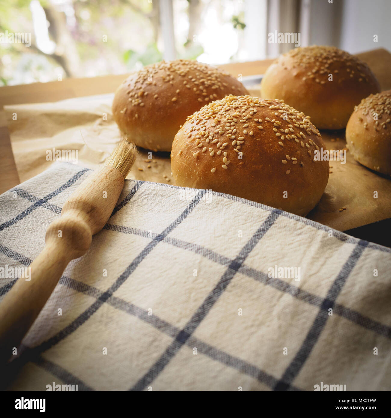 Top view of homemade sesame buns on a wooden chopping board with a striped white kitchen cloth and a pastry brush. Square format. Stock Photo