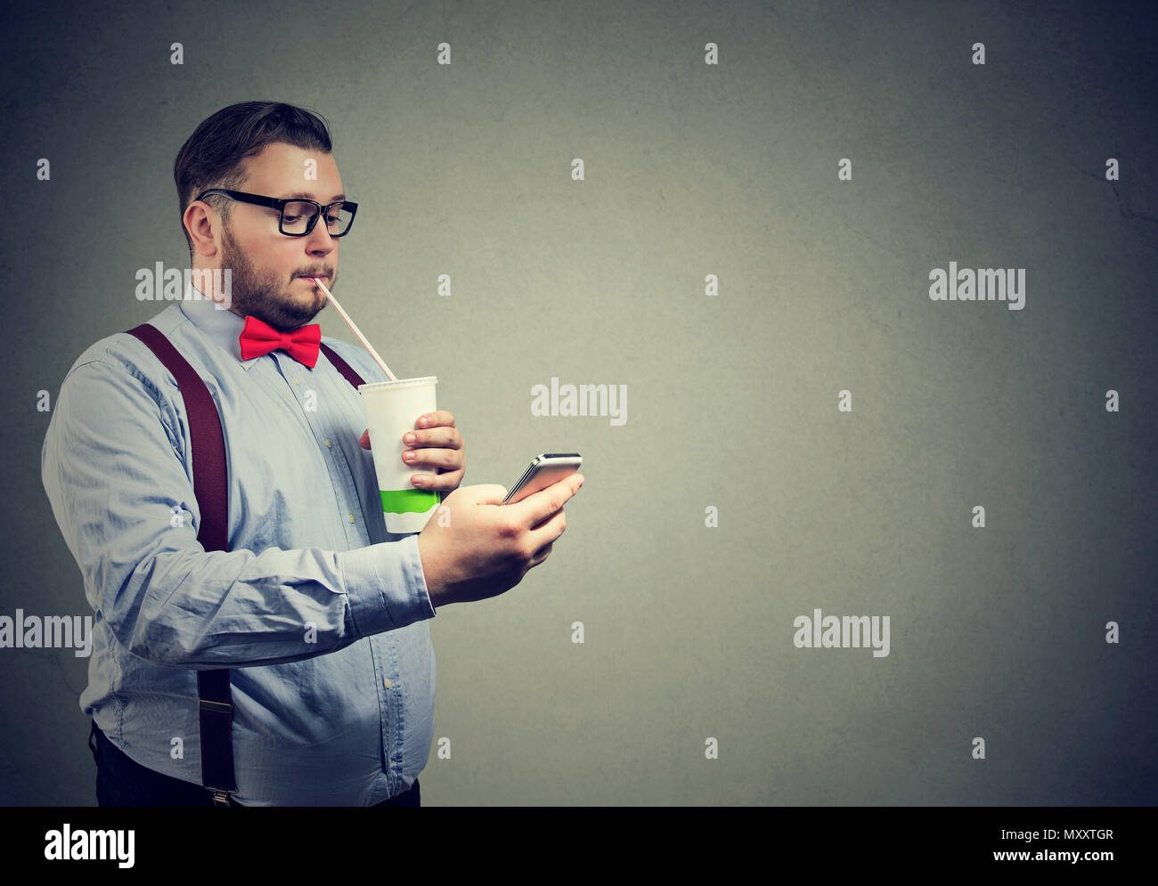 Young chunky man in formal outfit and glasses drinking sweet soda while using smartphone in leisure Stock Photo