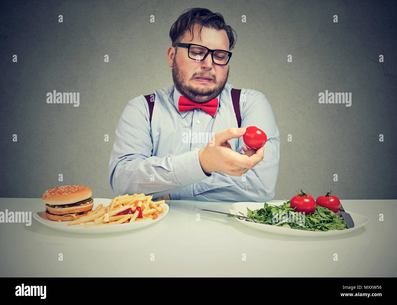 Overweight man in bow tie craving for burger and looking with disgust at salad and tomatoes sitting at table Stock Photo