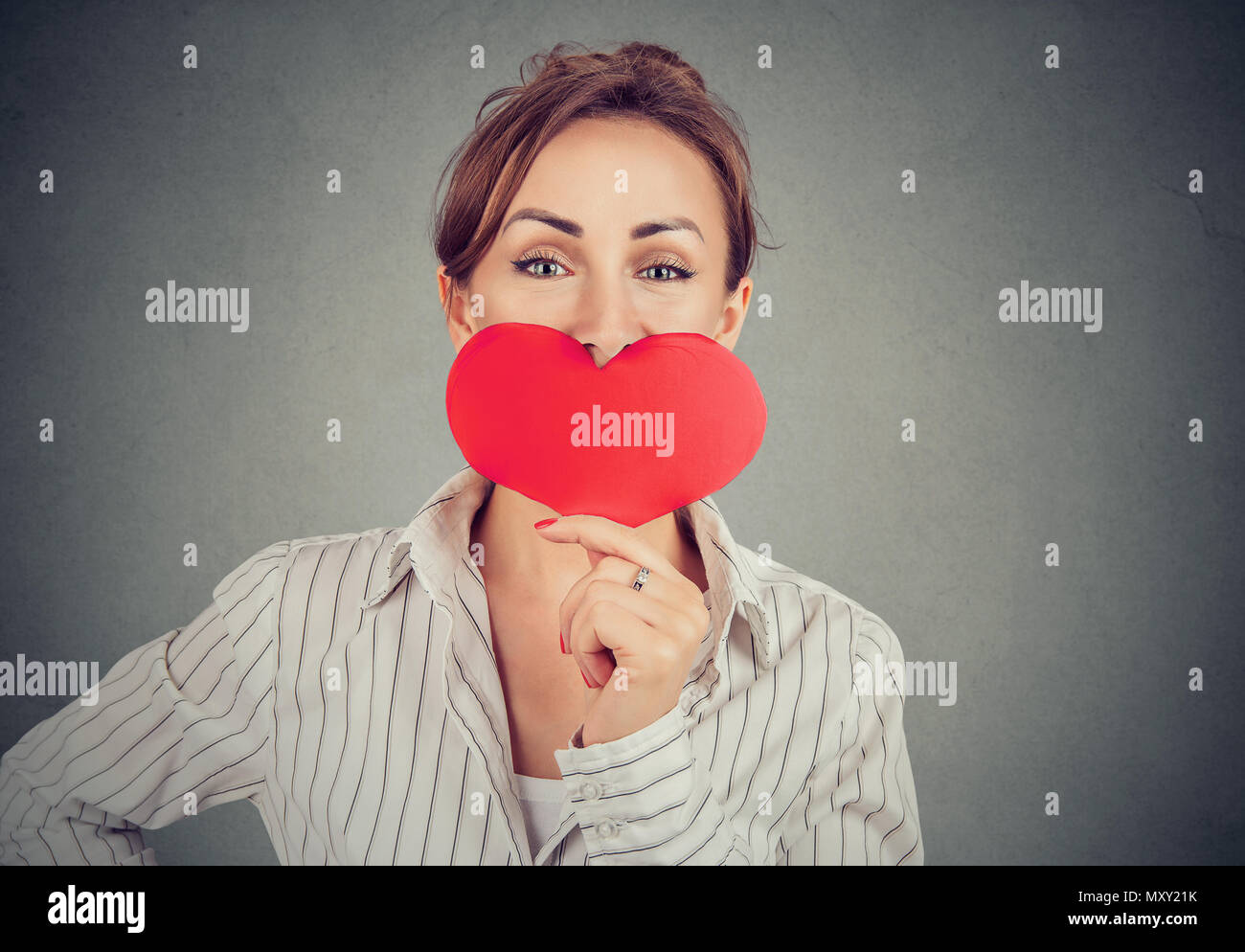 Beautiful positive girl covering mouth with big red paper heart and looking at camera with kind eyes on gray Stock Photo
