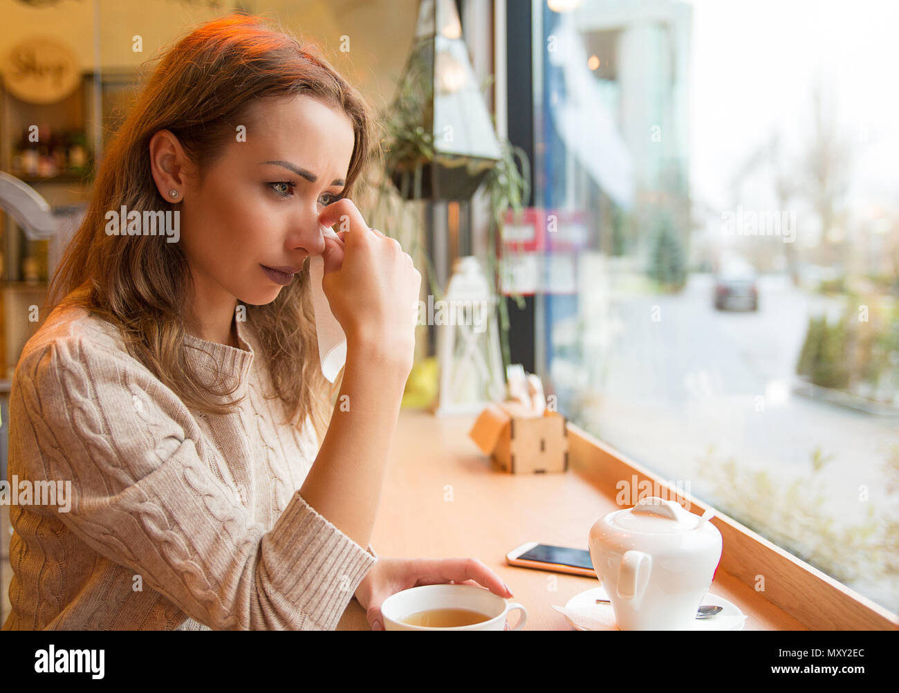 Young girl in sorrow crying after breakup sitting with cup of tea at table in cafeteria near window Stock Photo