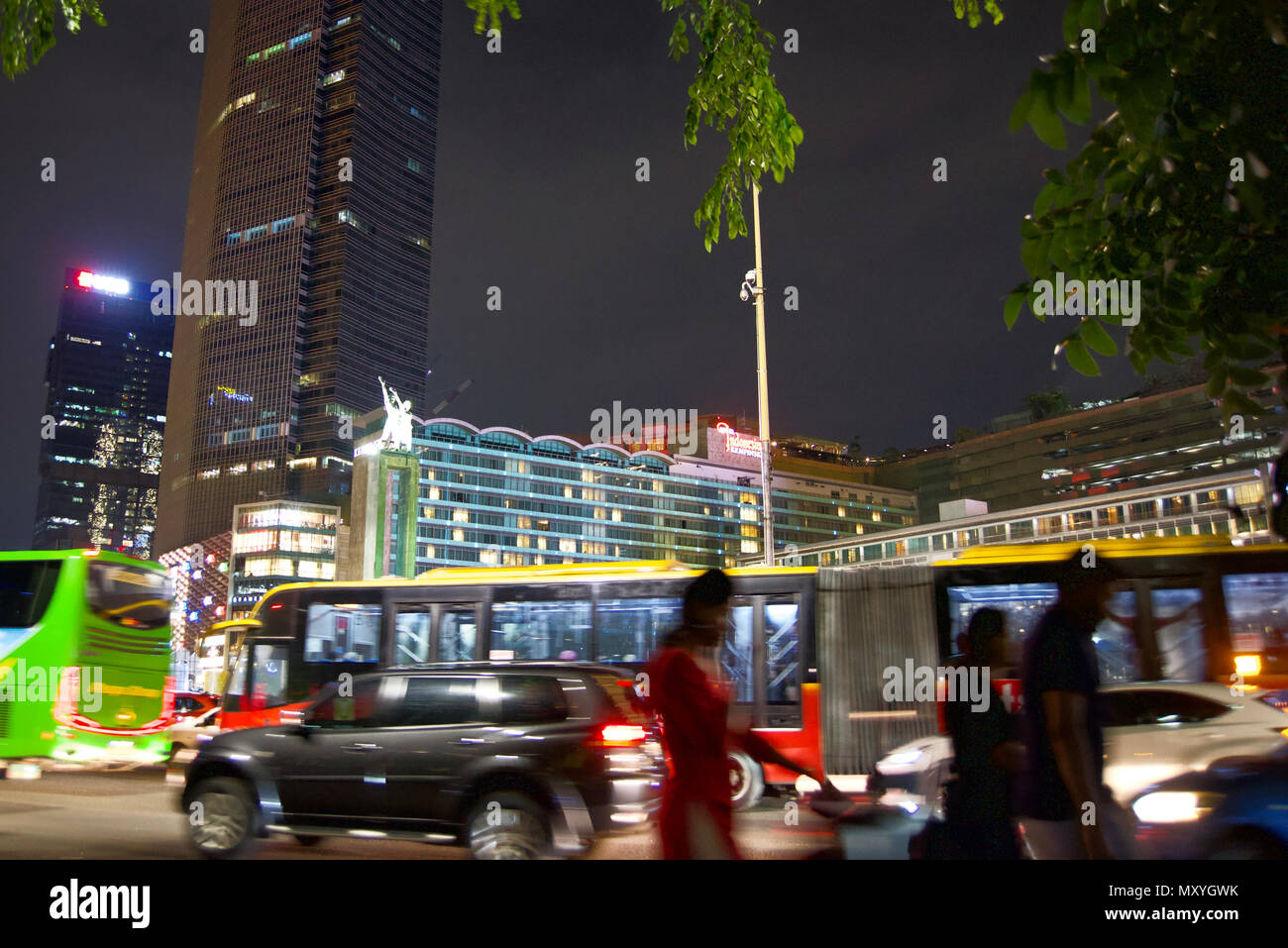 Pedestrians walk at night on roadside of Thamrin Street, Jakarta, with buses, cars, the welcome statue, and buildings in the background. Stock Photo