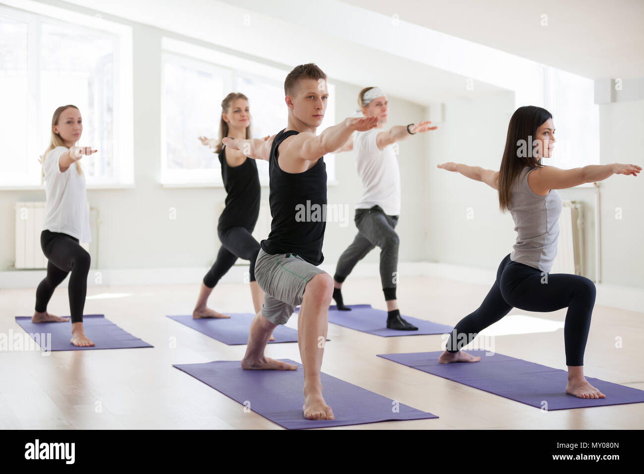 Group of young sporty people practicing yoga lesson, doing Warrior Two exercise, Virabhadrasana 2 pose, working out, indoor session full length, stude Stock Photo