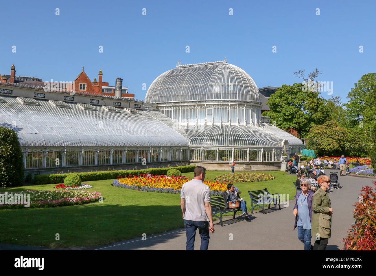 People in a UK park enjoying the sun and flowers beside the glass Palm House in Botanic Gardens, Belfast, Northern Ireland. Stock Photo